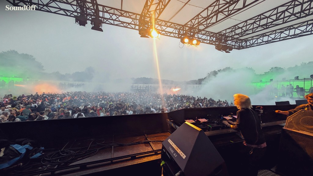 View from a festival DJ booth over a packed outdoor crowd, haze, and stage lights.